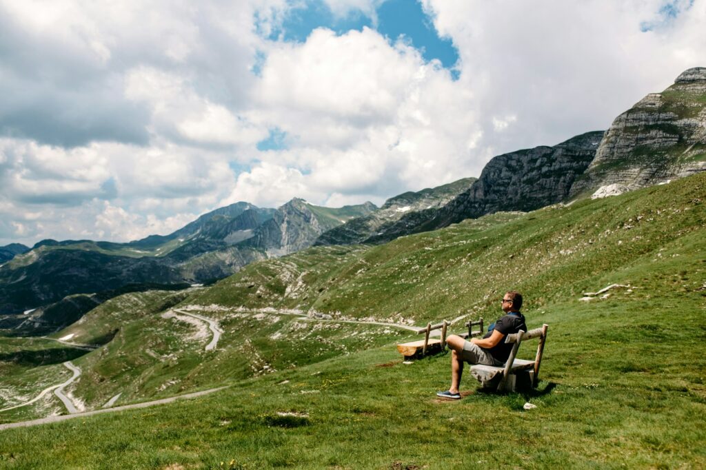 Single millennial man in sunglasses sits on a bench on a mountain plateau and looks at mountains